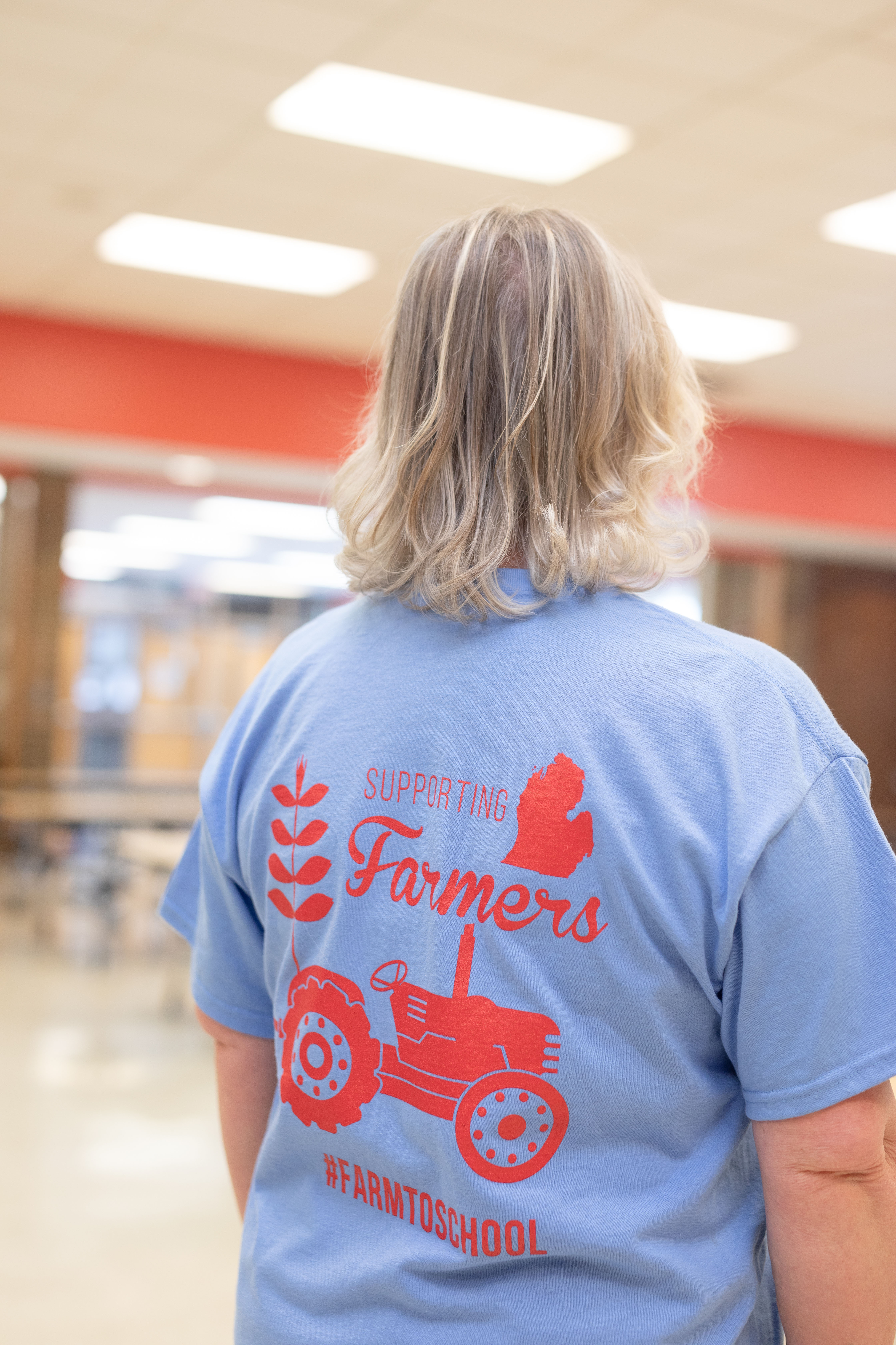 Person in a Michigan school cafeteria wears a farm to school shirt and faces away from the camera